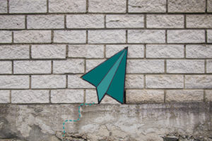 green paper plane by a white brick wall